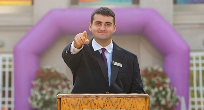 A confident male participant of the Leadership Development program stands poised at a podium, gesturing enthusiastically towards the vibrant crowd outside during the Best Buddies Leadership Conference.